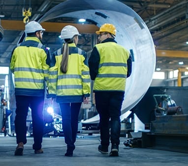 three people in safety jackets and hard hats walking through factory for company that uses it services